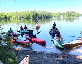 Several people in kayaks and two in a tandem canoe gather close together at a launch site in East Kingston. 